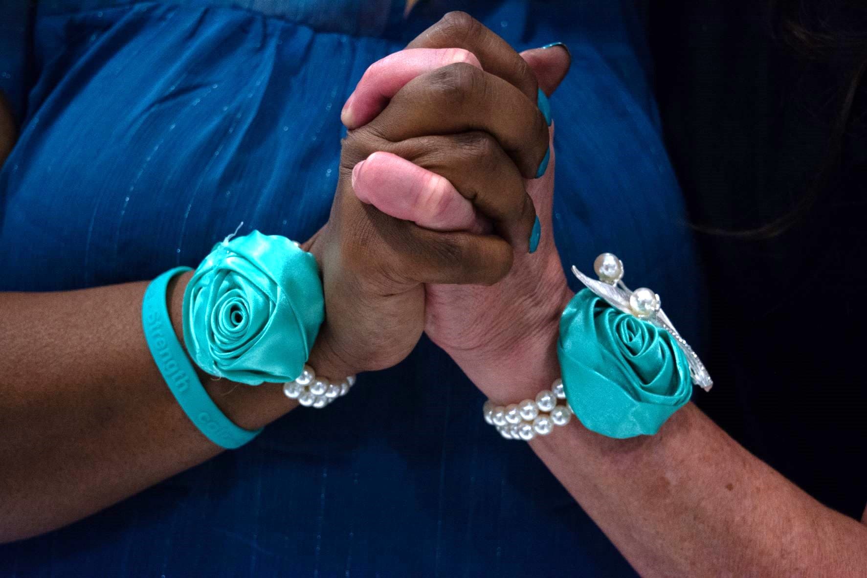 Two clasped hands, both wearing teal satin rose corsages and pearl bracelets